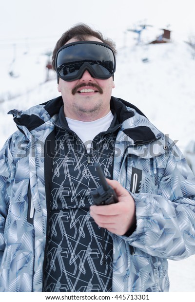 Half-length portrait of smiling man in black ski\
goggles holding radio standing\
outdoor.