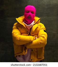 Half-length portrait of young anonymous man wearing pink balaclava and yellow down jacket, coat isolated on dark vintage background. Concept of safety, art, fashion.