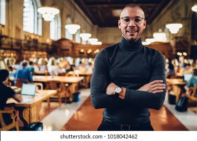 Half-length Portrait Of Young Prosperous Male Professor In Eyeglasses Spending Time In Public Library, Handsome Successful Writer Looking At Camera Standing With Crossed Arms Posing In Reading Hall