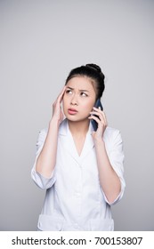 Half-length Shot Of Young Woman In Nurse Outfit Talking On The Phone, Looking Concerned And Touching Her Temple.