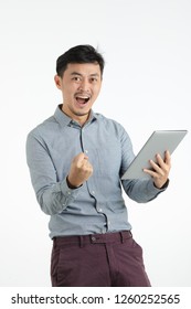 Half-length Studio Portrait Of Black Hair, Young Asian Man In Grey Shirt, Clenched Fist, Wide-open Mouth In Satisfied Gesture, Holding Tablet, Looking Straight To Camera, On Isolated White Background