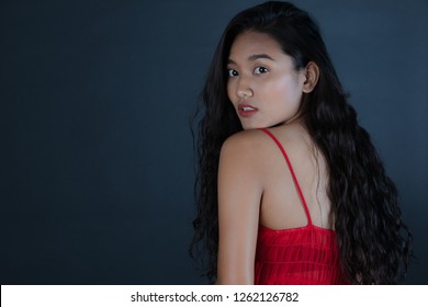 Half-length studio portrait of young beautiful,long,curly black hair Asian woman in red bare shoulder dress,turning back,tilted head,looking away from camera, on dark background