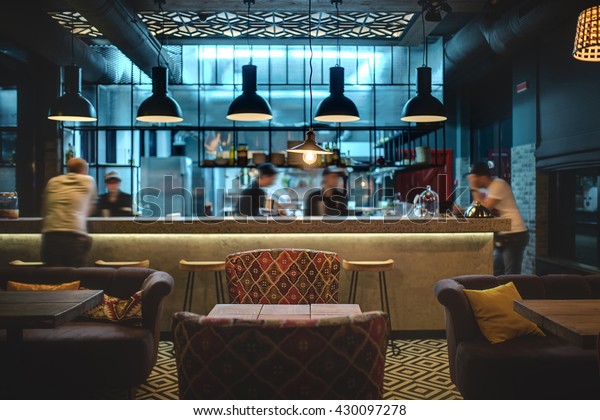 Half-lighted hall in a loft style in a mexican\
restaurant with open kitchen on the background. In front of the\
kitchen there are wooden tables with multi-colored chairs and\
sofas. On the sofas\
there