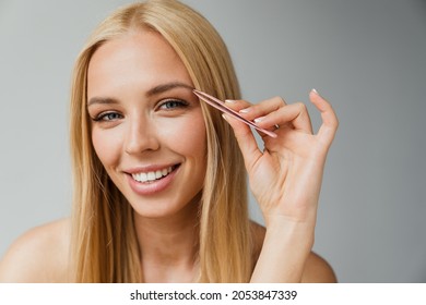 Half-naked blonde woman plucking her eyebrow with tweezers isolated over white background
