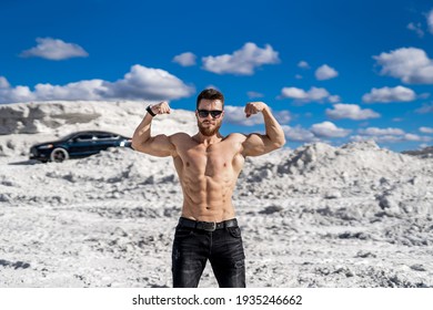 Halfnaked bodybuiler posing with muscular arms up. Background from beautiful white mountains sky and dark modern car. Man in suglasses standing.