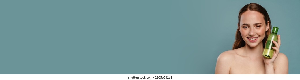 Half-naked Ginger Woman Smiling And Showing Face Lotion Isolated Over Blue Background