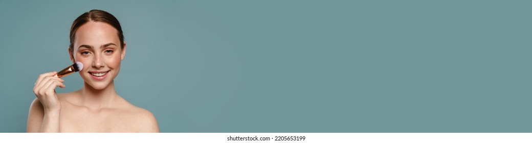 Half-naked Ginger Woman Smiling And Using Cosmetic Brush Isolated Over Blue Background