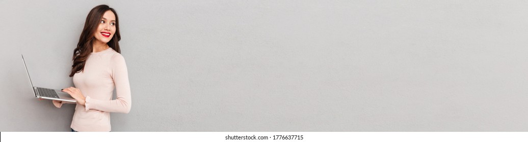 Half-turn image of cute female in casual clothes working on silver notebook, turning back over gray wall copy space