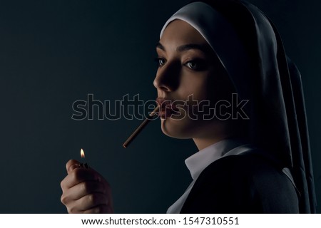 Half-turn portrait of a nun, taking on a black background. She wearing dark nun's clothing. The nun is lighting a cigarette. She looking to the left. 