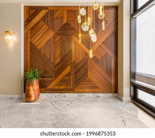 Hall with wood cladding wall decorated with geometrical patterns, tall rounded wooden planter with green bushes, contemporary glass tall chandelier, big window, and white marble floor in modern house