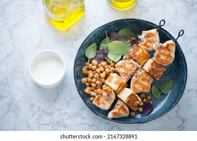 Halloumi and chicken kebabs with roasted chickpeas in a blue bowl, elevated view on a light-grey marble background, horizontal shot