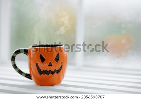 Halloween celebration concept with spooky jack lantern big mug at autumn season. All hallows eve party symbol hot beverage in orange pumpkin mug on wooden windowsill at window background