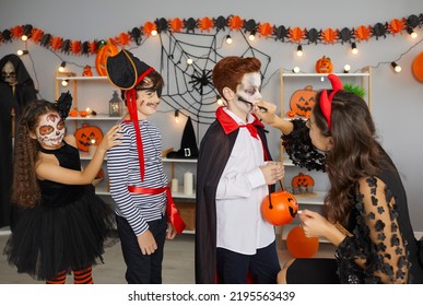 Halloween Children's Makeup. Young Woman Prepares Children For Costume Party And Applies Makeup To Little Boy Making Him Vampire. Boys And Girls Have Fun In Room With Halloween Decor.