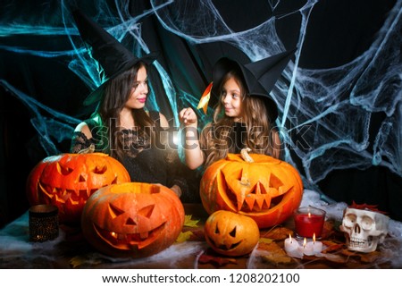 Halloween Concept - beautiful caucasian mother and her daughter in witch costumes enjoy using magic with magic wand to halloween pumpkin jar over spider web on black studio background.