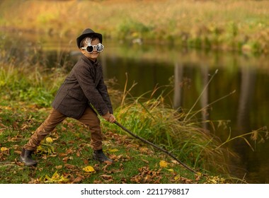 Halloween Concept: Cute Little 5 Yo Child (boy) In Skeleton Glasses,warm Brown Coat (jacket), Hat, Boots Holds Stick, Stands Near Pond And Scaring People In Autumn Yellow Park, Fall Season. Horizontal