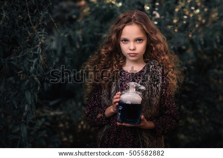 Halloween Concept! Cute little witch with a bottle of blue  smoking potion in the decorated garden.  Beautiful child girl in witch costume  with halloween decoration.