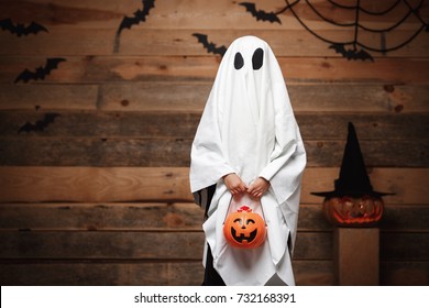 Halloween Concept - Little White Ghost With Halloween Pumpkin Candy Jar Doing Trick Or Treat With Curved Pumpkins Over Bats And Spider Web On Wooden Studio Background.