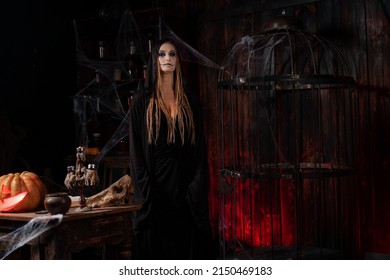 Halloween Concept. Witch Dressed Black Hood With Dreadlocks Standing Dark Dungeon Room Use Magic Book For Conjuring Magic Spell. Female Necromancer Wizard Gothic Interior With Skull, Cage, Spider Web