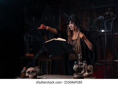 Halloween Concept. Witch Dressed Black Hood Standing Dark Dungeon Room Use Magic Book Put Her Hand Forward Conjuring Magic Spell. Female Necromancer Wizard Gothic Interior With Skull, Cage, Spider Web
