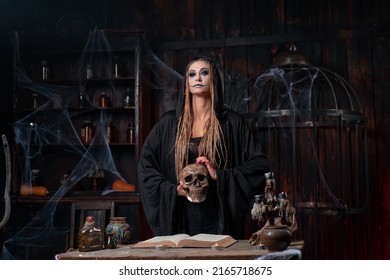 Halloween Concept. Witch Dressed Black Hood Holding Skull In Hand Standing Dark Dungeon Room Use Magic Book For Conjuring Magic Spell. Female Necromancer Wizard Gothic Interior