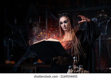 Halloween Concept. Witch Dressed Black Hood Standing Dark Dungeon Room Use Magic Book Conjuring Magic Spell. Fire Sparkles Magic Female Necromancer Wizard Gothic Interior
