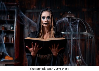 Halloween concept. Witch dressed black hood with dreadlocks standing dark dungeon room use magic book for conjuring magic spell. Female necromancer wizard gothic interior looking camera medium shoot - Shutterstock ID 2169554459