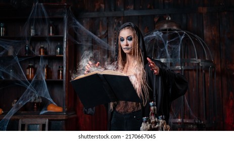Halloween Concept. Witch Dressed Black Hood Standing Dark Dungeon Room Use Levitating Magic Book Conjuring Magic Spell. Female Necromancer Wizard. Smoking Spell Book