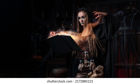 Halloween Concept. Witch Dressed Black Hood With Dreadlocks Standing Dark Dungeon Room Use Magic Book For Conjuring Magic Spell. Female Necromancer Wizard Gothic Interior Sparkles Soul Effect