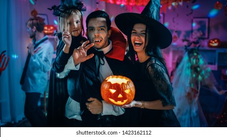 Halloween Costume Party: Father Count Dracula Holds Little Bat Girl Daughter And Hugs Witch Wife For A Happy Family Portrait. In The Background Monsters Having Fun