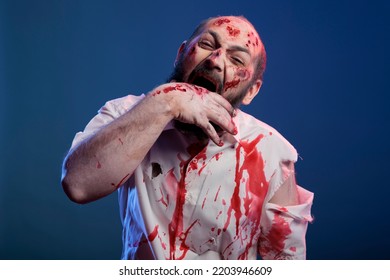 Halloween Cruel Zombie Biting Hand With Scars And Bloody Wounds, Standing In Studio. Apocalyptic Frightening Corpse And Brain Eating Villain With Deadly Horror Face And Aggressive Sinister Eyes.