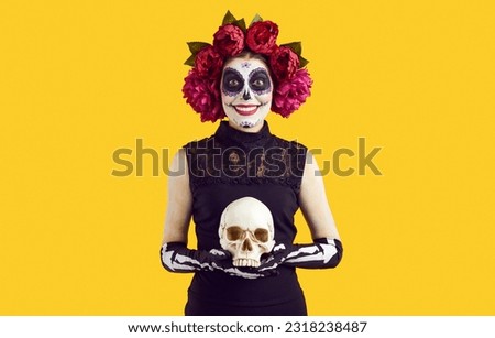 Halloween or Day of the Dead. Cheerful woman with mexican style face art posing with decorative skull on orange background in studio. Woman with bright makeup and peony wreath smiles at camera.