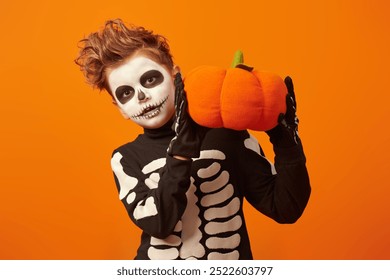 Halloween and The Day of the Dead. Cute little boy with makeup and in a costume of skeleton holds a toy pumpkin and smiles. Orange studio background with copy space. - Powered by Shutterstock