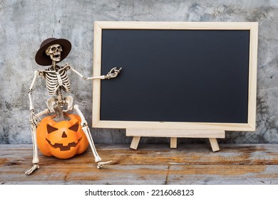 Halloween decoration. Funny human skeleton wearing a hat sitting on a pumpkin head Jack-o'-lantern with blackboard background copy space leave blank for writing greetings. - Powered by Shutterstock