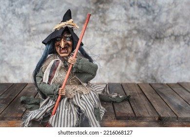 Halloween Decoration. Old Woman Witch Doll With Scary Face Holding A Broom Sitting On An Old Wooden Slat With Dark Background And Copy Space Bare Plaster Or Loft Style.