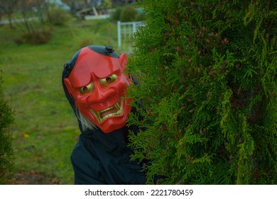 Halloween Devil Mask For A Woman, A Woman Behind A Bush Near A House In A Garden. A Festive Event. Zina, Latvia - 02.11.2022