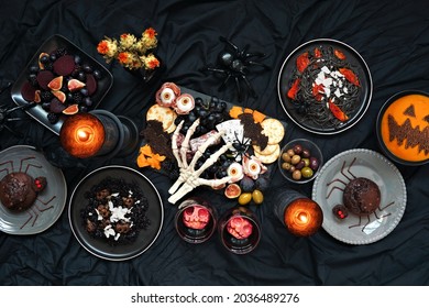 Halloween Dinner Party Table Scene Over A Dark Black Background. Top Down View. Scary Charcuterie Board, Black Risotto And Pasta, Spider Cakes, Jack O Lantern Pumpkin Soup And Apple Skull Wine