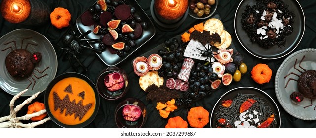 Halloween Dinner Party Table Scene Over A Dark Black Banner Background. Overhead View. Charcuterie Board, Black Risotto And Pasta, Spider Cakes, Jack O Lantern Pumpkin Soup And Apple Skull Wine.