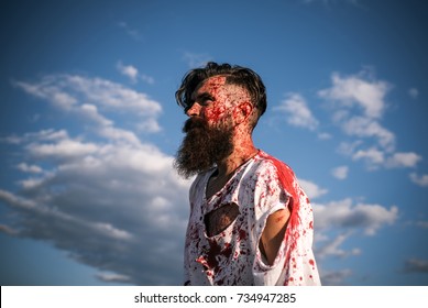 Halloween evil and horror. Injury, hurt, pain. Man with red blood splatters on bearded face. Zombie hipster in torn tshirt with bloodstains on blue sky. War soldier or victim concept.