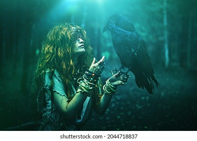 Halloween. A fairytale forest witch with a mask covering her eyes talks to a black raven, standing in the thicket of the forest. Mystical ritual of death. Woman shaman in ritual garment.