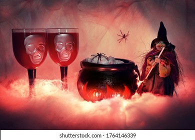 Halloween figure of a witch with a broom on a lighted smoky background of cobwebs, standing next to a burning witch's cauldron filled with food and snacks, on which a spider crawls, next to two creepy