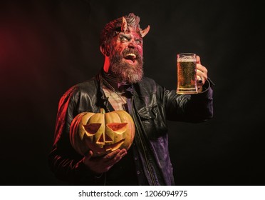 Halloween holiday celebration and party. Man with satan horns hold pumpkin. Devil smile with glass mug. Hipster demon drink beer on black background. Bad habits and addiction concept.