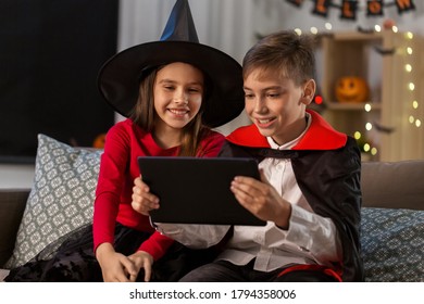 halloween, holiday and childhood concept - smiling boy and girl in party costumes of witch and dracula with tablet pc computer at home