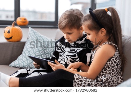halloween, holiday and childhood concept - smiling little boy and girl in party costumes with tablet pc computer at home