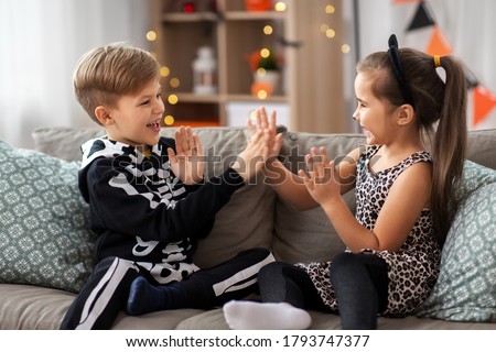 halloween, holiday and childhood concept - smiling little boy and girl in costumes playing clapping game at home