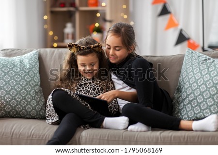 halloween, holiday and childhood concept - smiling little girls in costumes with tablet pc computer at home
