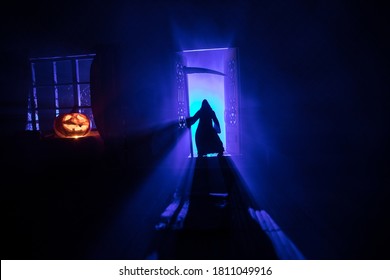 Halloween Horror Concept With Glowing Pumpkin. A Realistic Dollhouse Living Room With Furniture, Door And Window At Night. Grim Reaper On Door With Blade. Selective Focus.