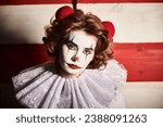 Halloween horrors. A scary sad clown girl with red hair, dressed in a white circus dress, looks at the camera while standing over a striped wall. 