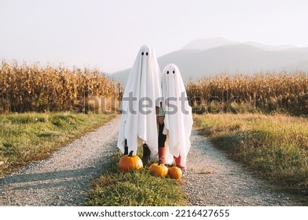 Halloween Kids Holidays Concept. Little children dressed in white sheets like as cute ghosts at countryside at fall season. Photo of white ghosts with orange pumpkins in cornfield background.