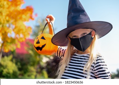 Halloween Kids Mask. Portrait Blonde Girl In Witch Costume With Pumpkin Bucket. Child Wearing Black Face Masks Outdoors Protecting From COVID-19.