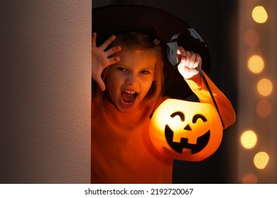 Halloween. Little girl in witch costume hides and scares from corner with lantern pumpkin. Child in black hat plays in horror stories in dark
